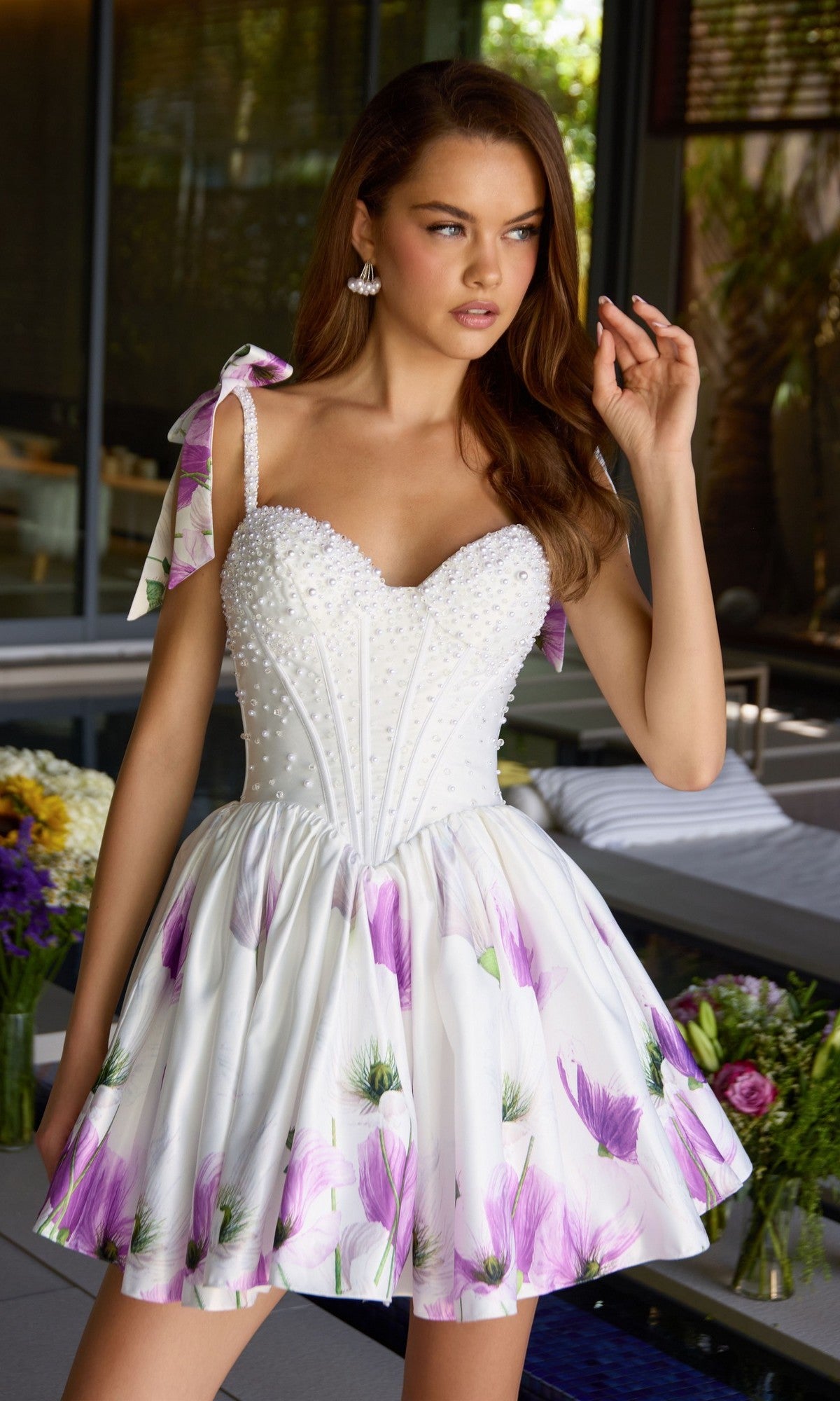 A woman in the Ava Presley 42432 short cocktail dress, featuring a pearl-embellished corset and flared floral satin skirt, stands indoors by a flower-adorned table, gazing thoughtfully to the side.